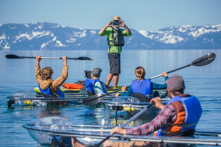 People kayaking in clear kayaks with snow-capped mountains in the background.