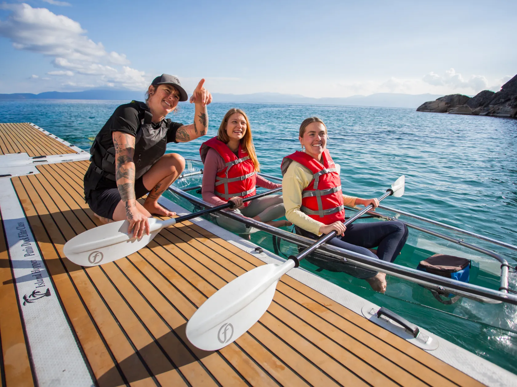 Three people in life vests on a dock with paddles, near a clear kayak in the water.