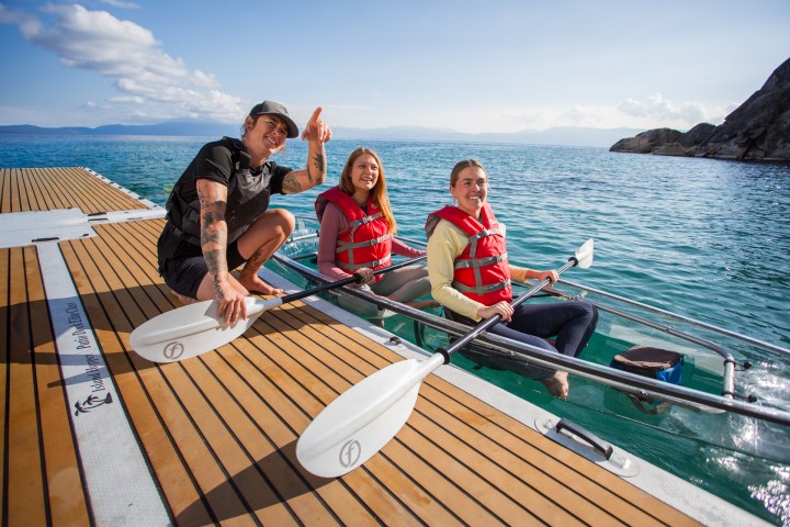 Three people in life vests on a dock with paddles, near a clear kayak in the water.