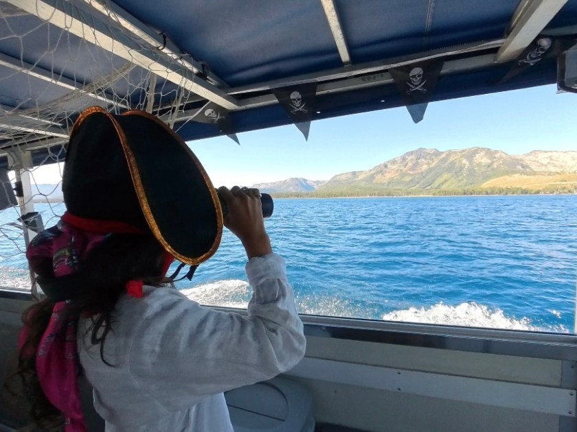 Person in pirate costume using binoculars on a boat with mountains in the background.