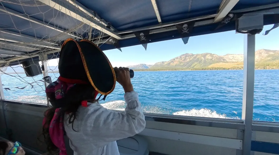 Screenshot Person in pirate costume using binoculars on a boat with mountains in the background.