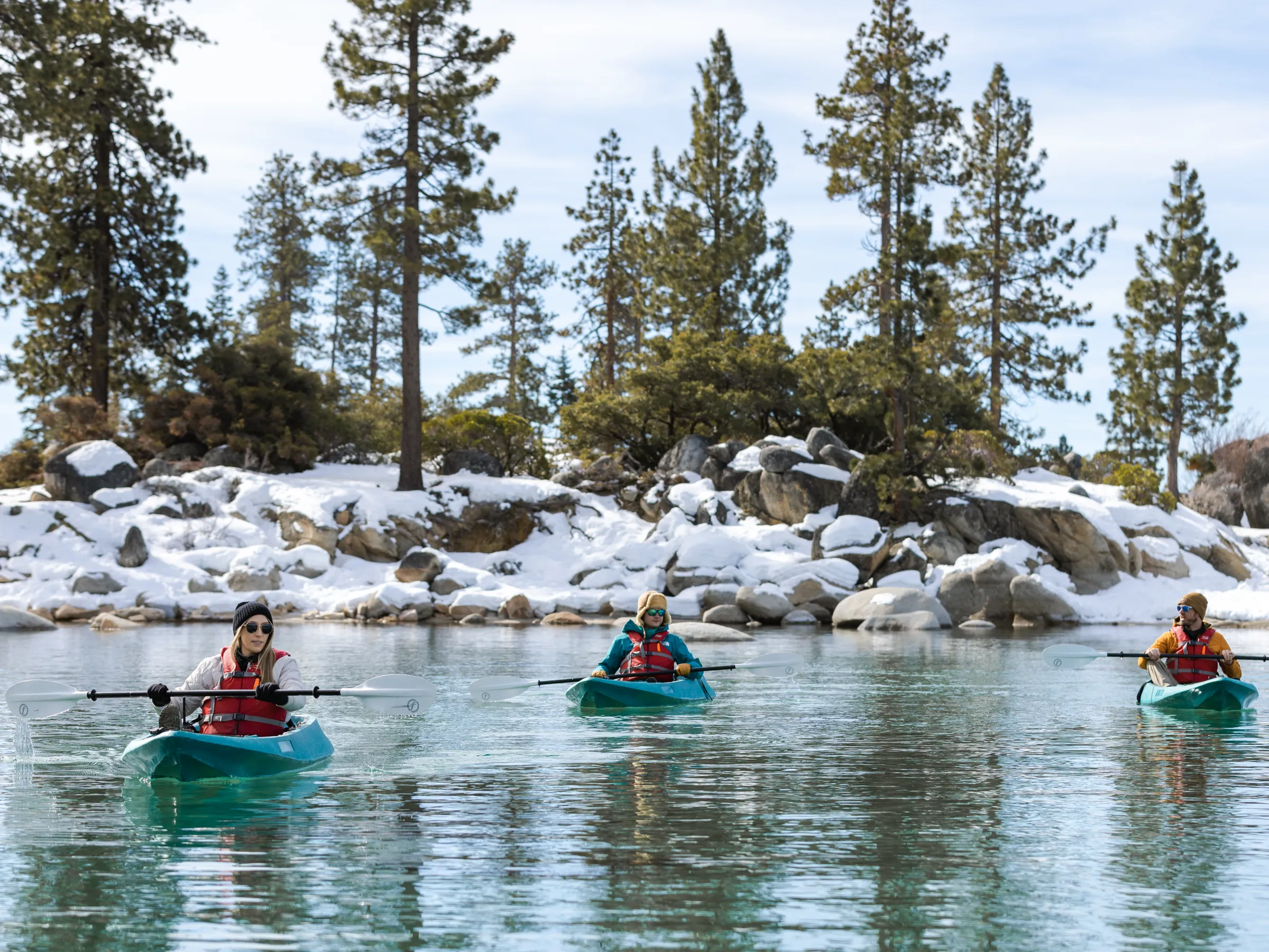 Three kayakers paddle on a lake surrounded by snow-covered rocks and pine trees.