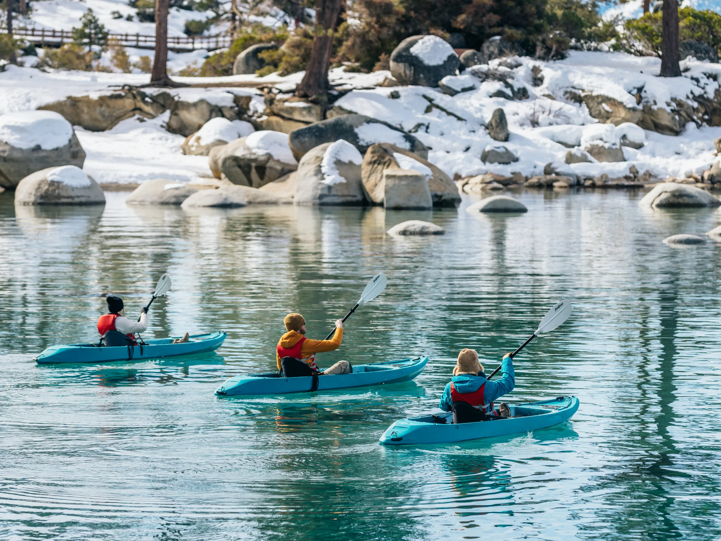 Three people kayaking on a tranquil lake surrounded by snow-covered rocks and trees.