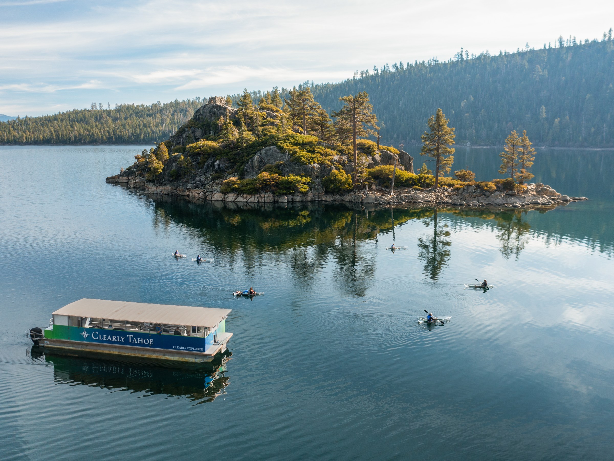 People kayaking on a lake near a wooded island, with a tour boat in the foreground.