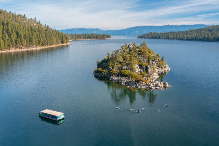 Aerial view of a lake with a small island, surrounded by mountains and trees, with a boat and kayakers nearby.