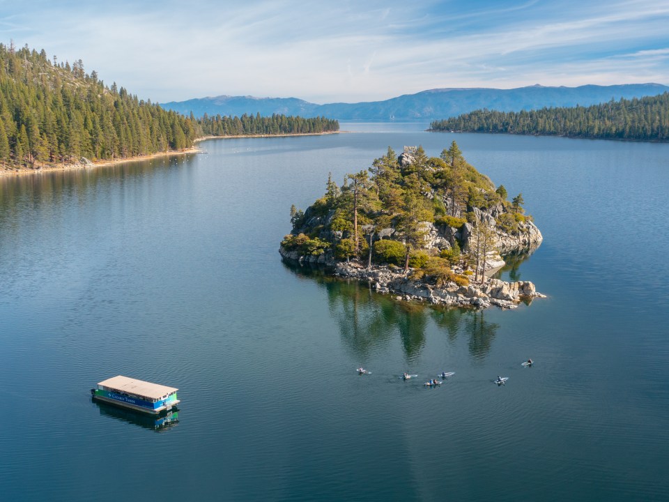 Aerial view of a lake with a small island, surrounded by mountains and trees, with a boat and kayakers nearby.
