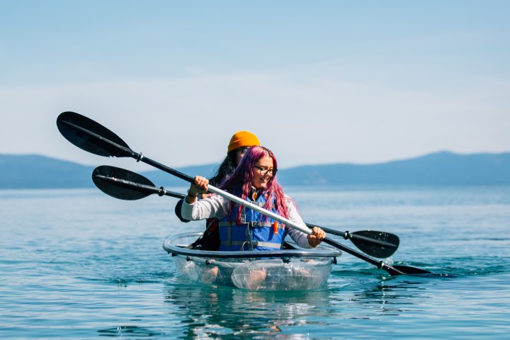 Two people in a clear kayak paddling on a calm blue lake under a clear sky.