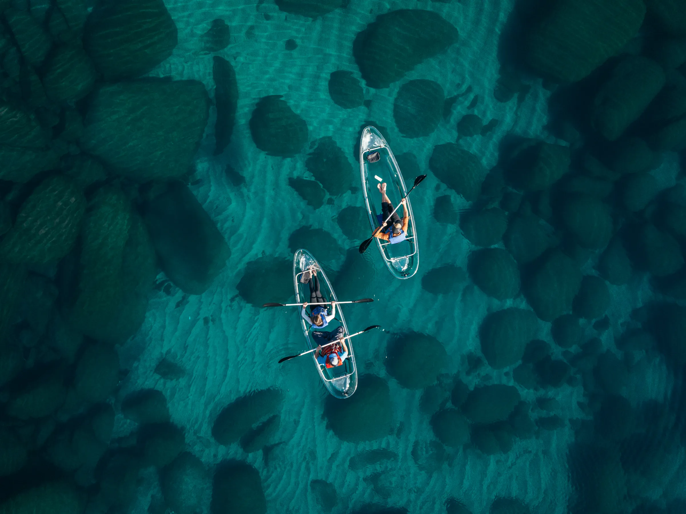 Two people kayaking in transparent boats over clear blue water with visible rocks.