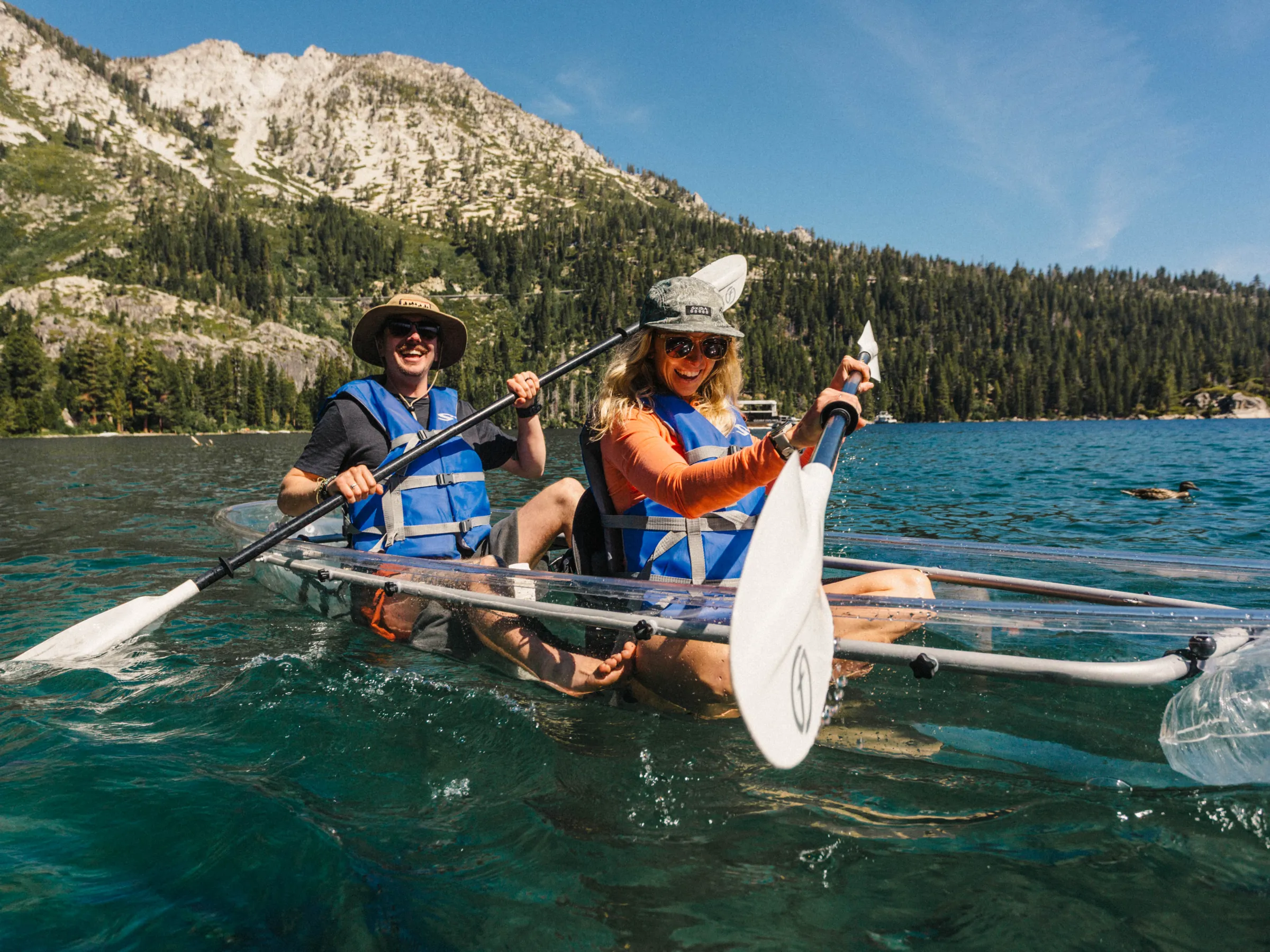 Two people kayaking in clear blue water with mountains and forest in the background.
