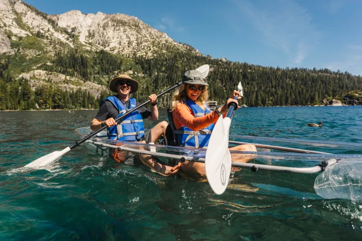 Two people kayaking in clear blue water with mountains and forest in the background.