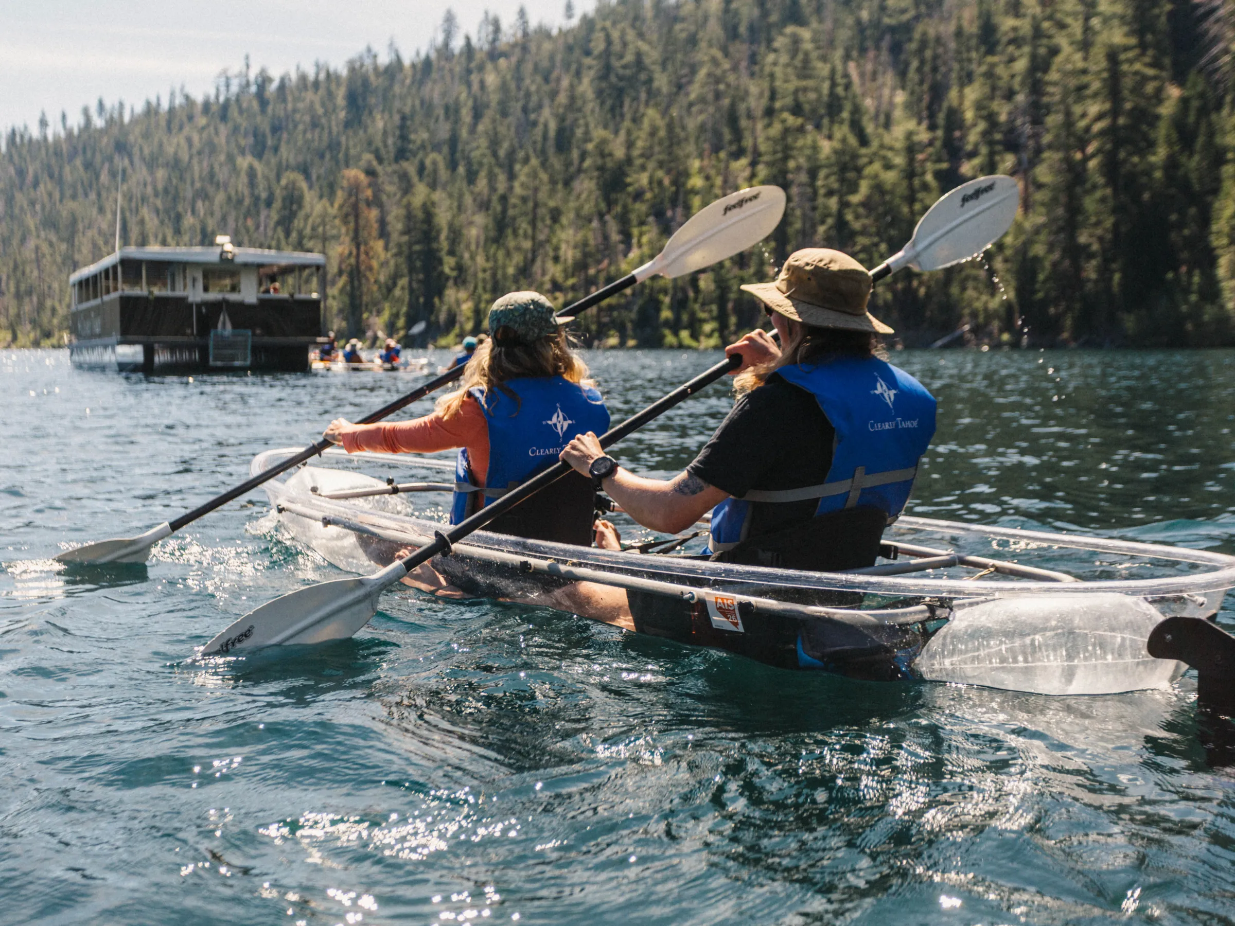 Two people kayaking on a clear blue lake with a forest and boat in the background.