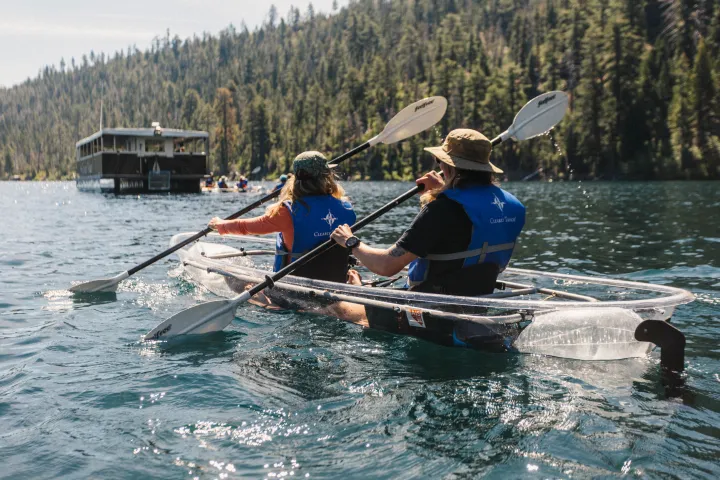 Two people kayaking on a clear blue lake with a forest and boat in the background.