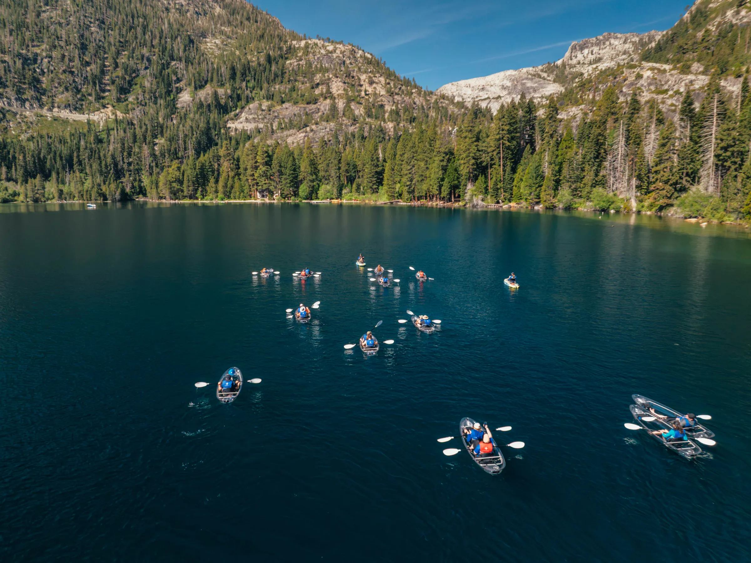 Groups of kayakers paddle on a lake surrounded by mountains and forest.