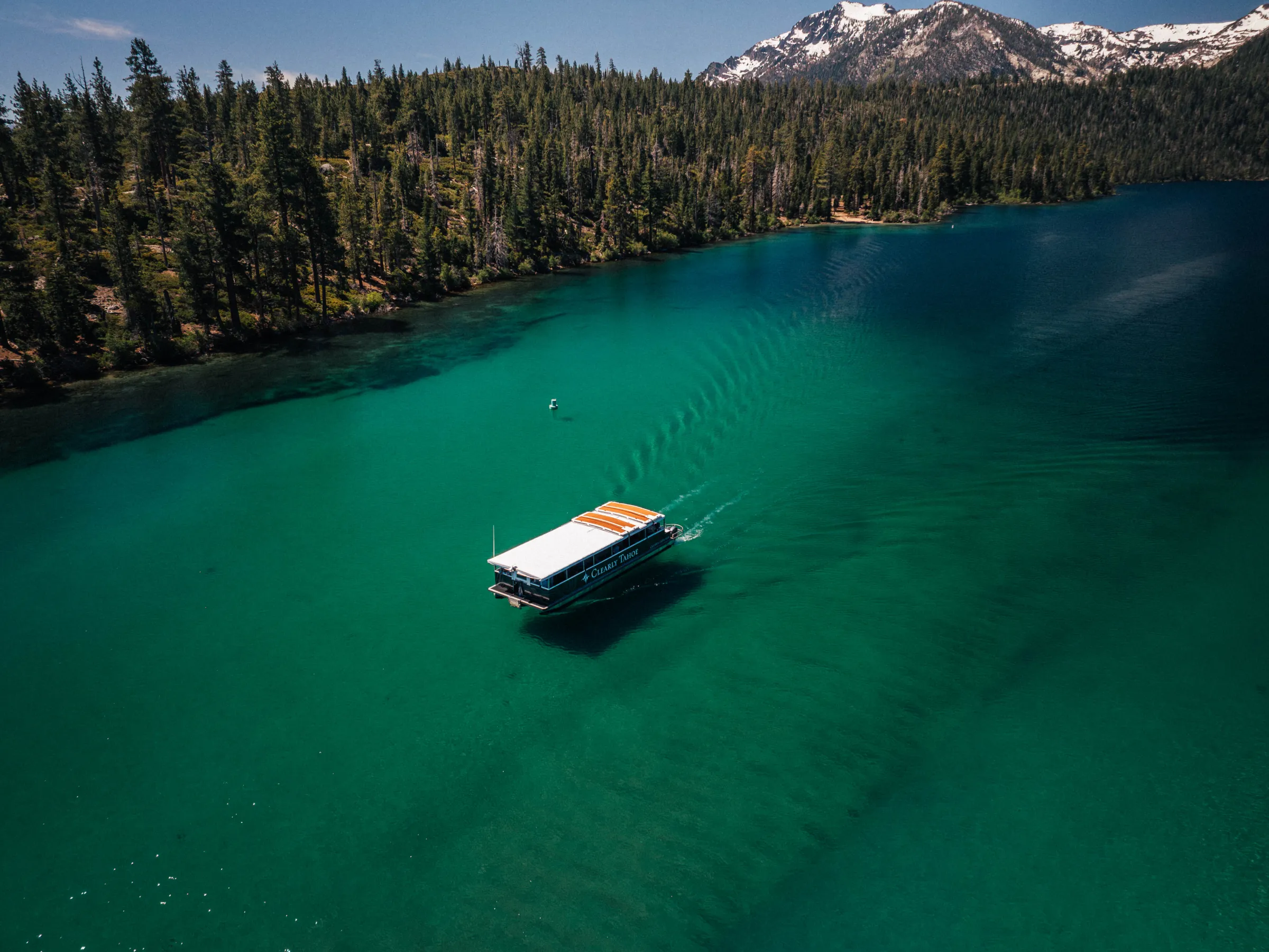 Boat on clear lake surrounded by pine trees and mountains, under blue sky.