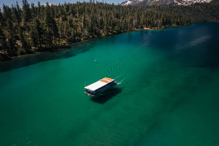 Boat on clear lake surrounded by pine trees and mountains, under blue sky.