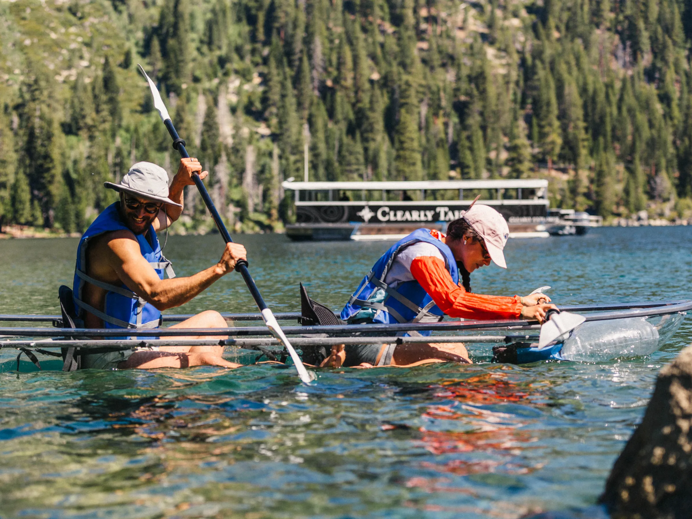 Two people kayaking on a clear lake with forested hills in the background.