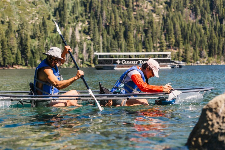 Two people kayaking on a clear lake with forested hills in the background.