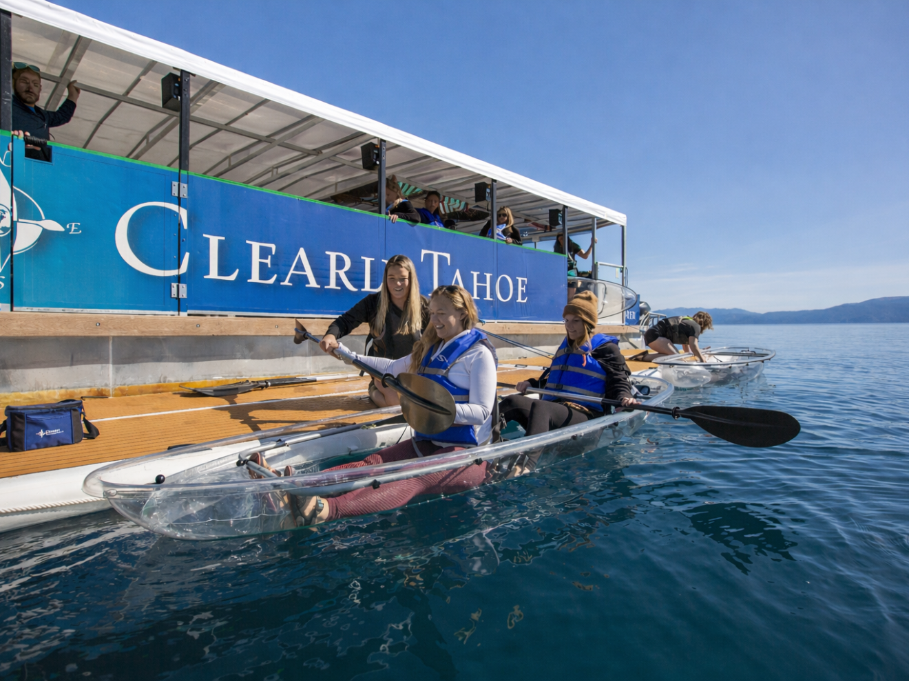 People in clear kayaks on a lake next to a boat with 'Clear Lake Tahoe' sign.