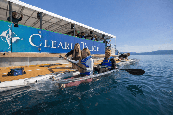 People in clear kayaks on a lake next to a boat with 'Clear Lake Tahoe' sign.