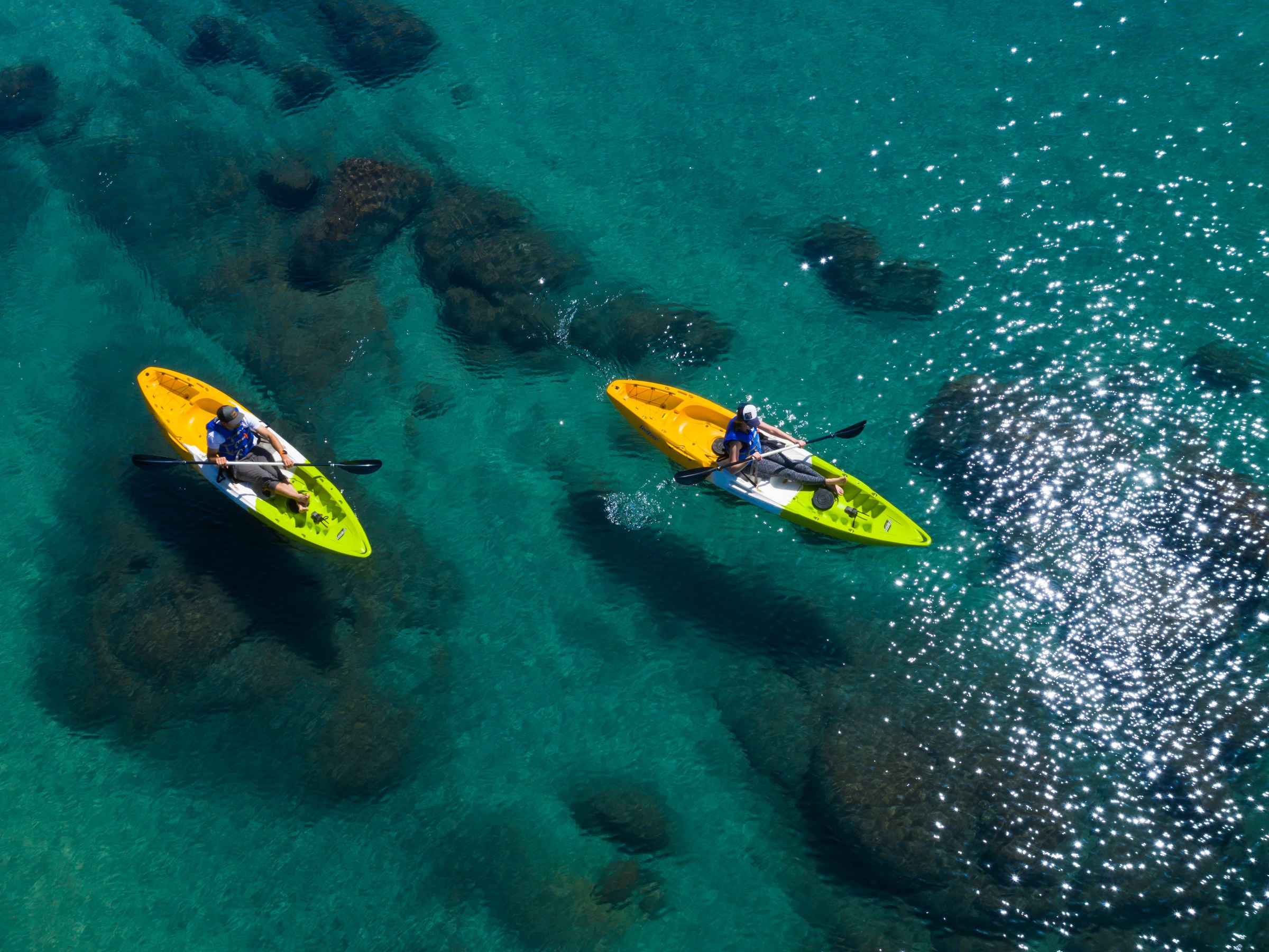 Two people kayaking on clear blue water with visible rocks.
