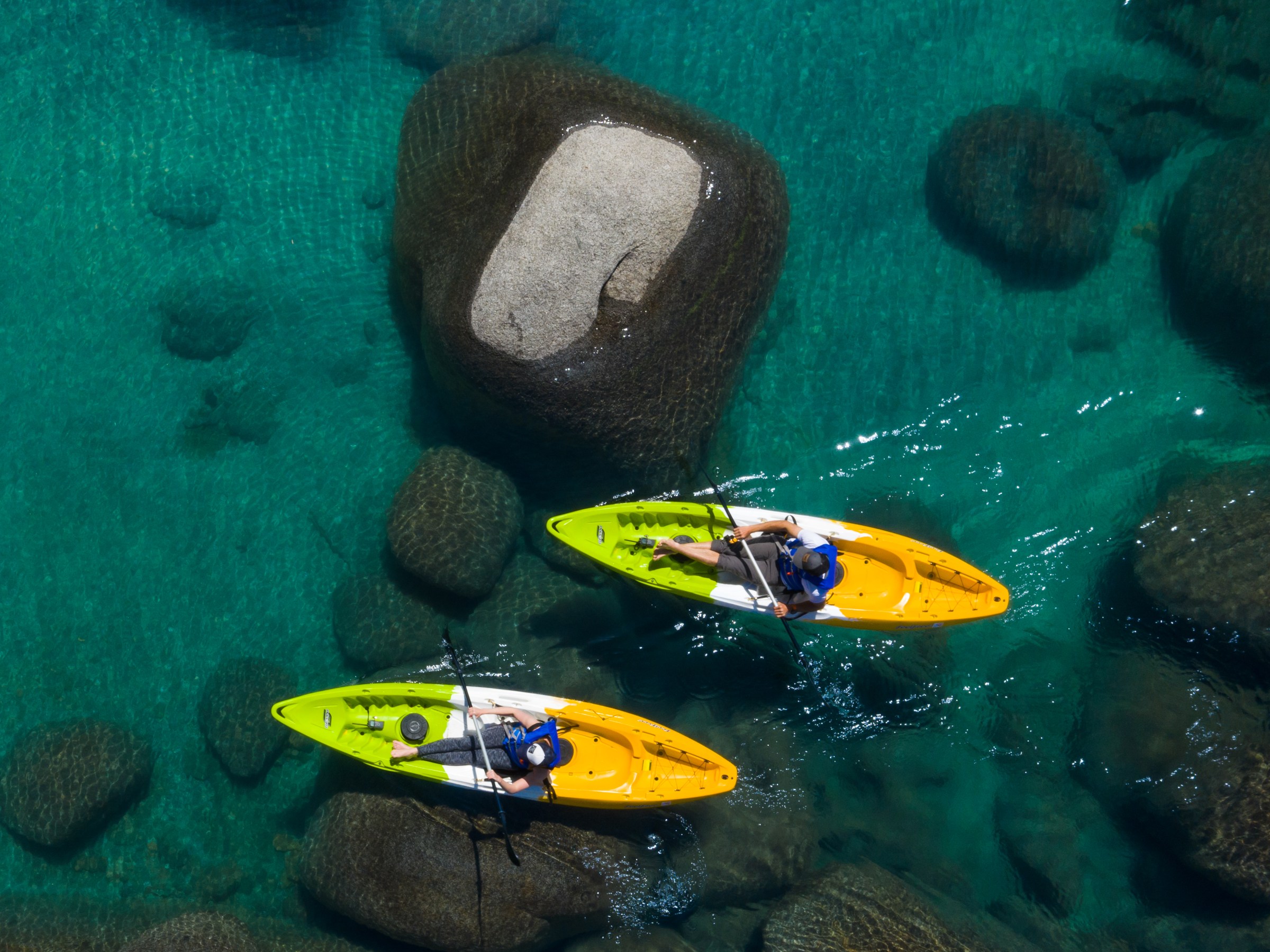 Two people kayaking on clear turquoise water with large rocks visible underneath.