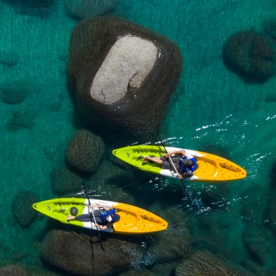 Two people kayaking on clear turquoise water with large rocks visible underneath.
