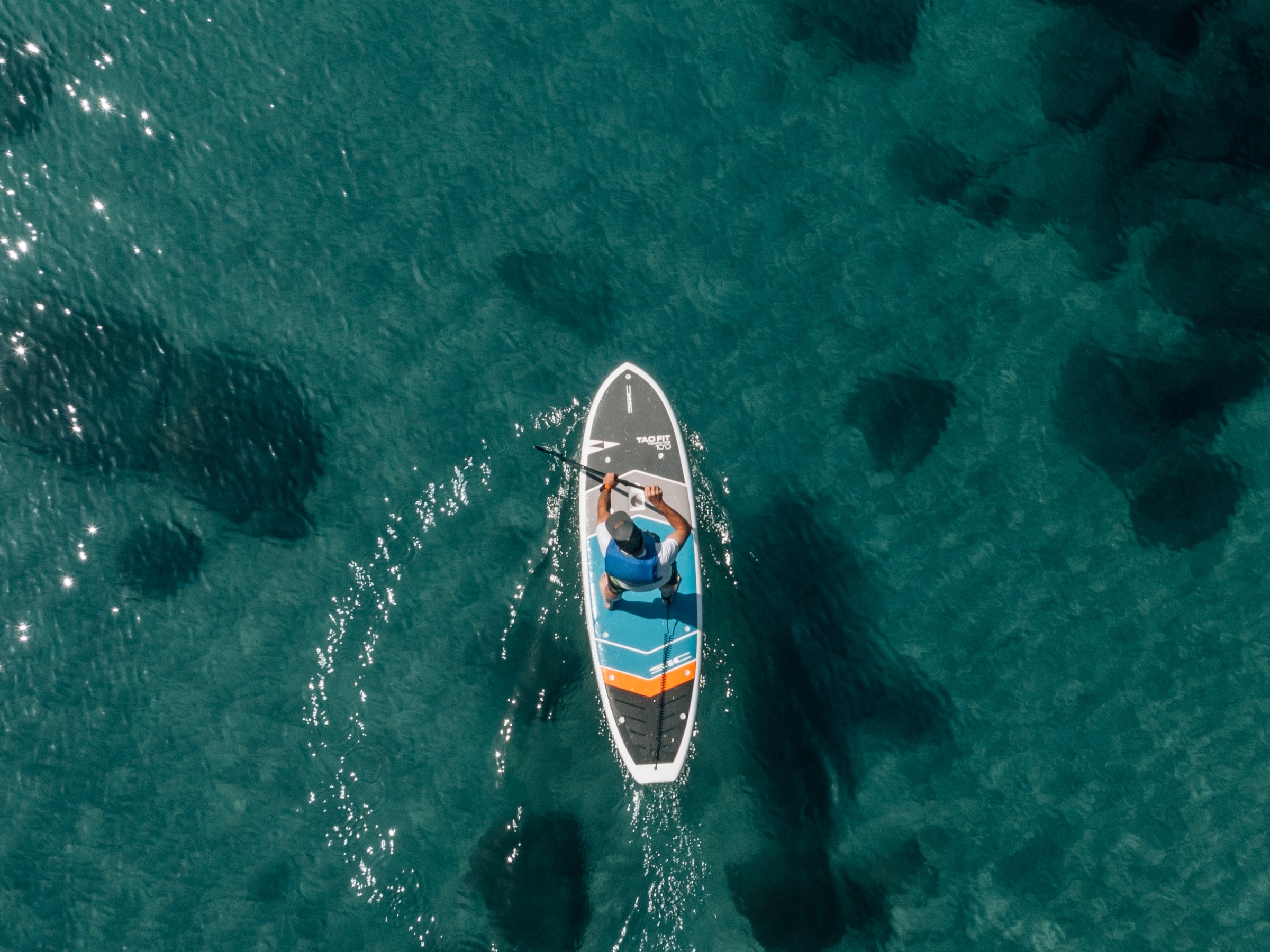 Person paddleboarding on clear turquoise water with visible underwater rocks.