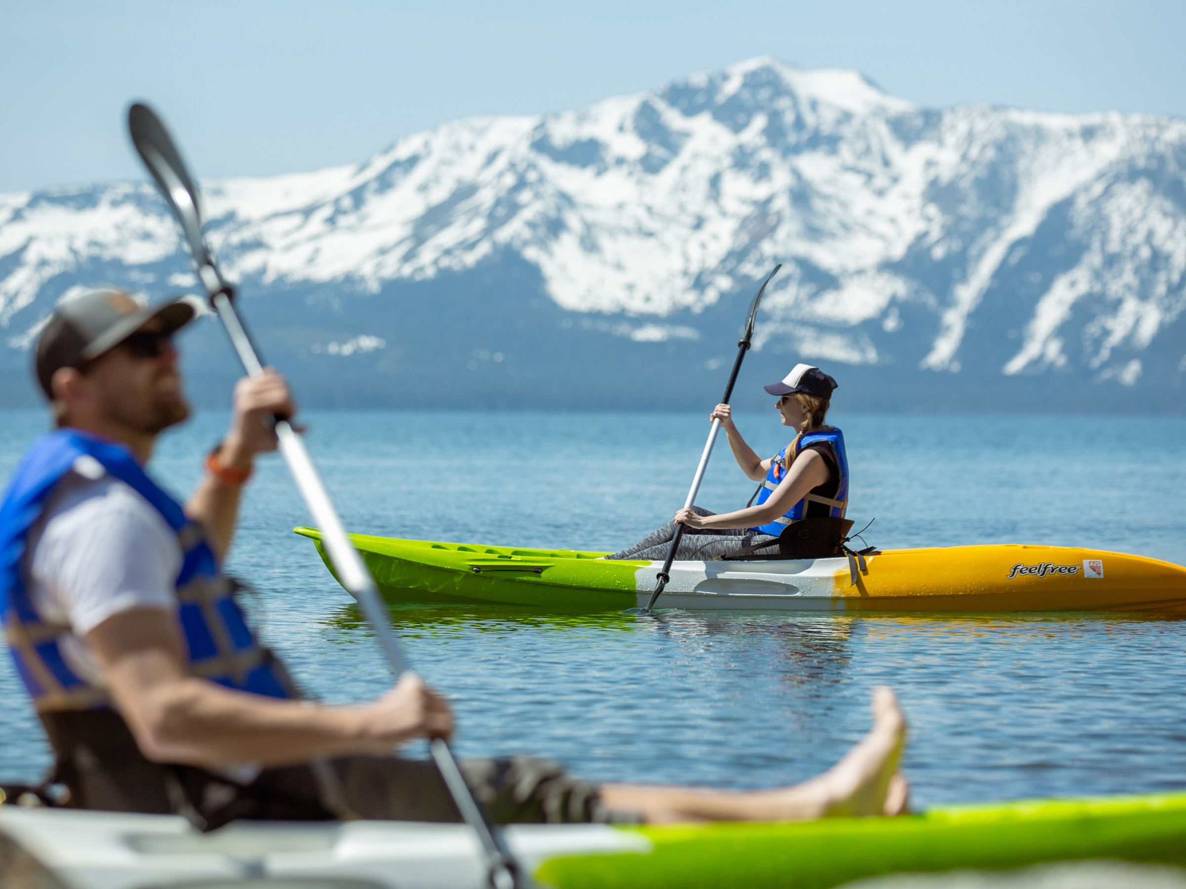 Two people kayaking on a lake with snow-capped mountains in the background.