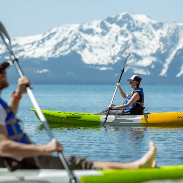 Two people kayaking on a lake with snow-capped mountains in the background.