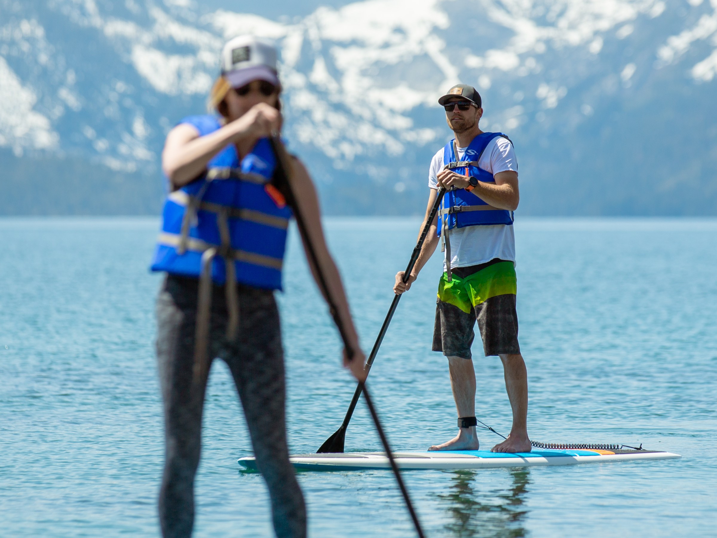 Two people paddleboarding on a lake with snowy mountains in the background.