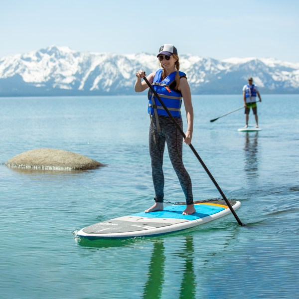 Two people paddleboarding on a calm lake with snow-capped mountains in the background.