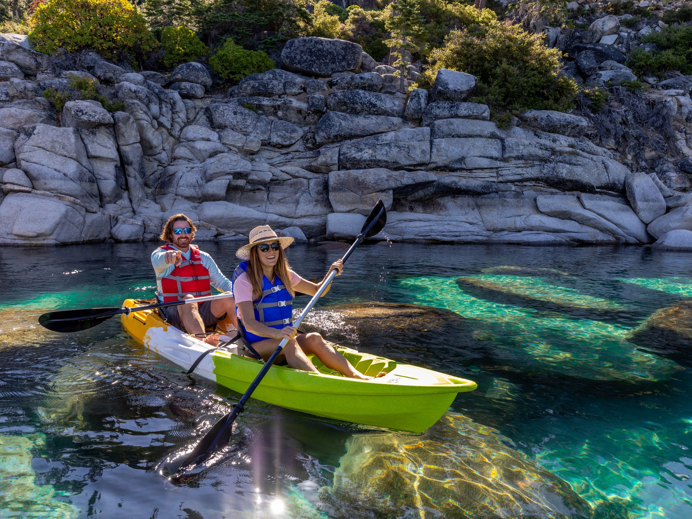 Two people kayaking on clear water near rocky shoreline.