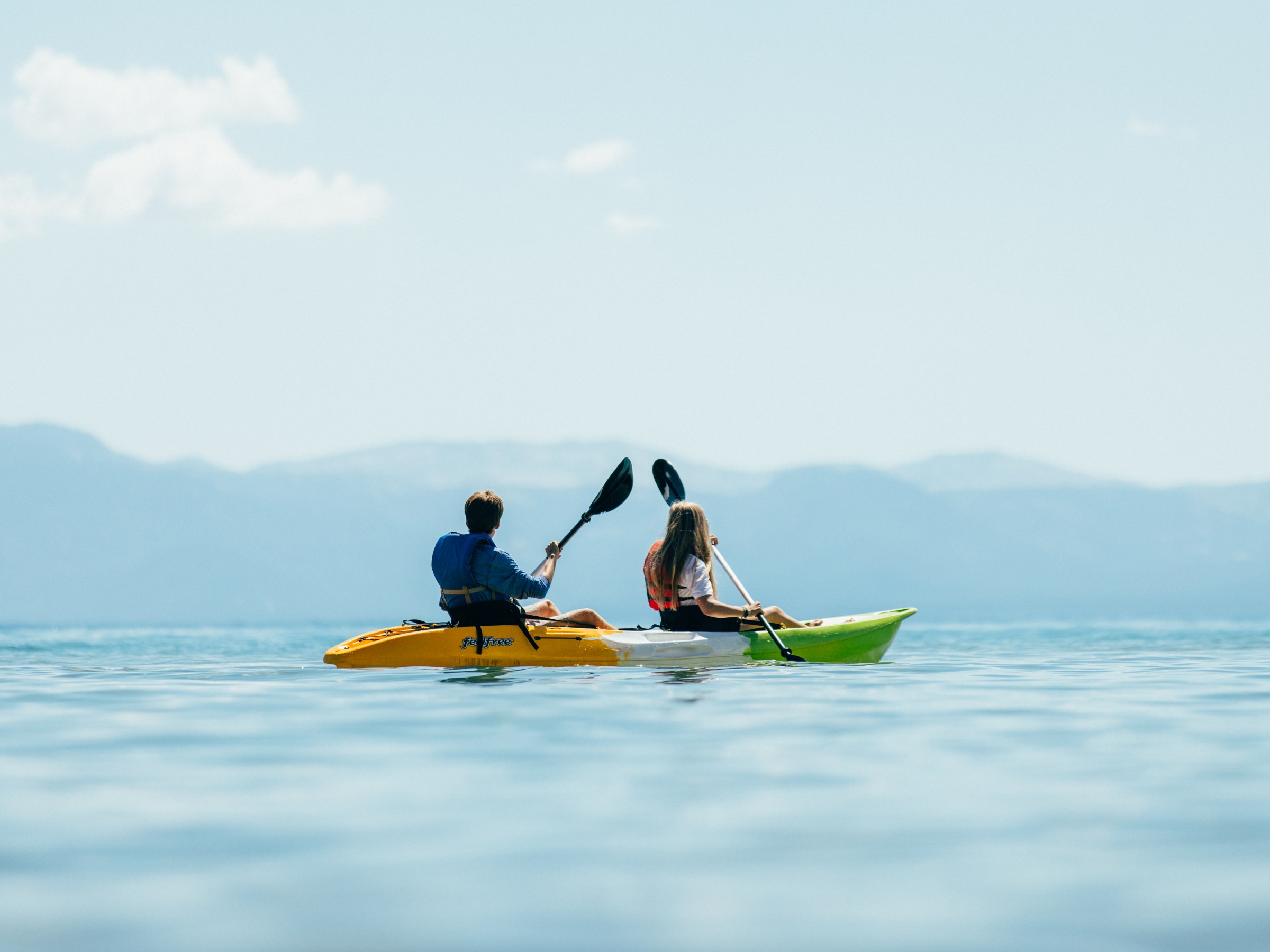 Two people kayaking on a calm lake with mountains in the background.