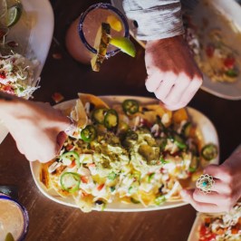 Hands grabbing loaded nachos from a platter on a wooden table.