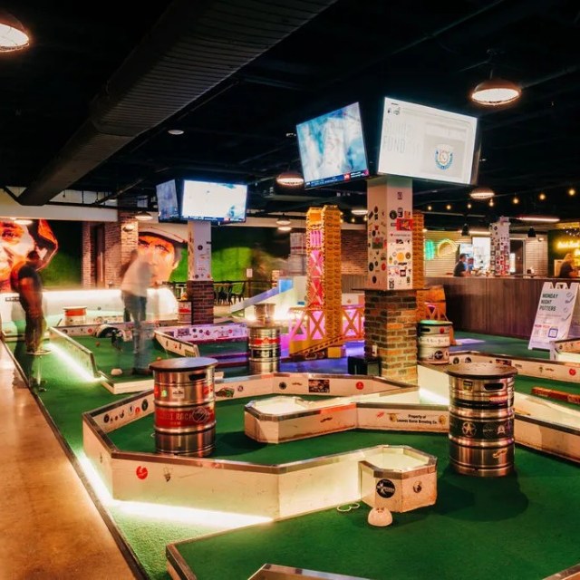 Indoor mini-golf course with colorful decor and overhead screens.