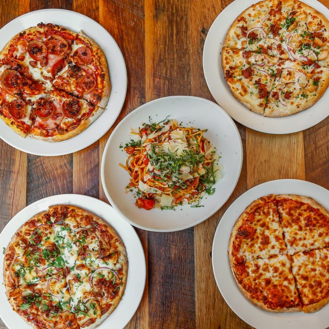 Four pizzas and a pasta dish on wooden tables.
