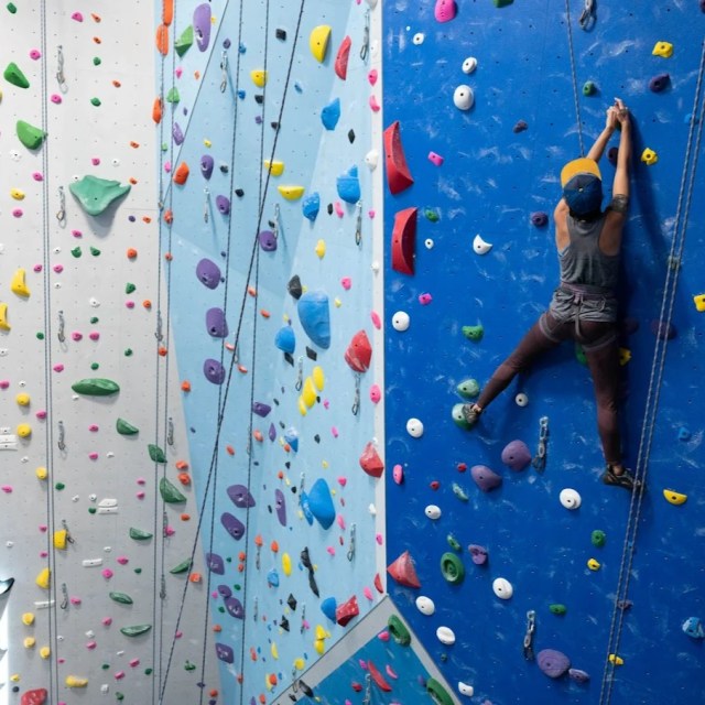 Person rock climbing indoors on a colorful climbing wall.