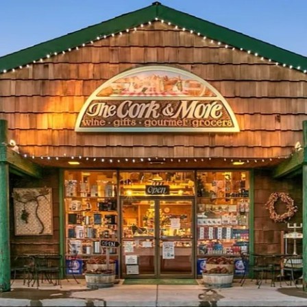 Front view of a decorated wooden store named 'The Cork & More' with lights and outdoor seating.