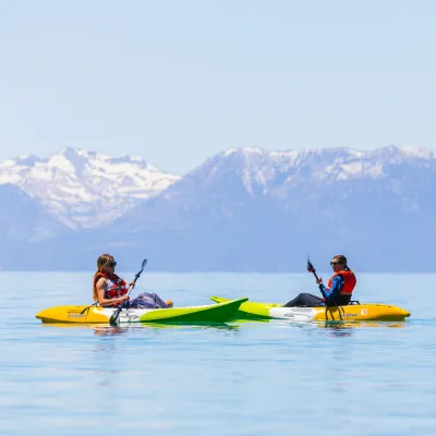 Two people kayaking on a lake with snow-capped mountains in the background.