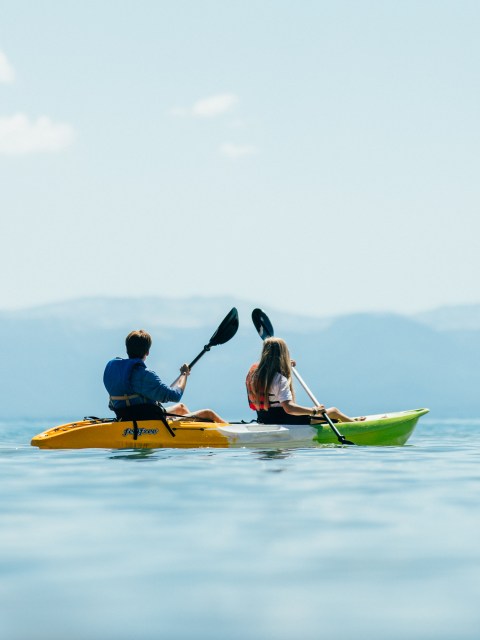 Two people kayaking on a calm lake with mountains in the background.