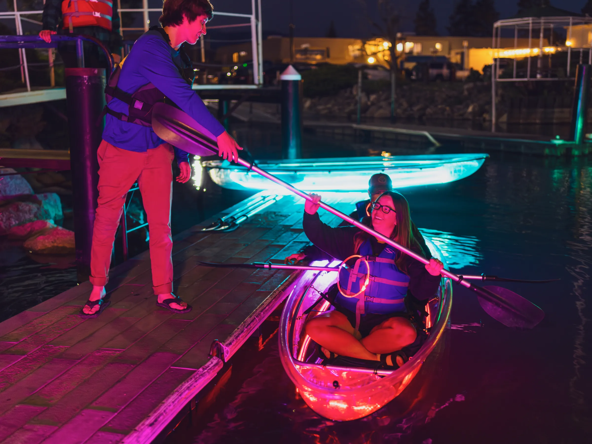 Person on dock helps another in a kayak at night with colorful lights.