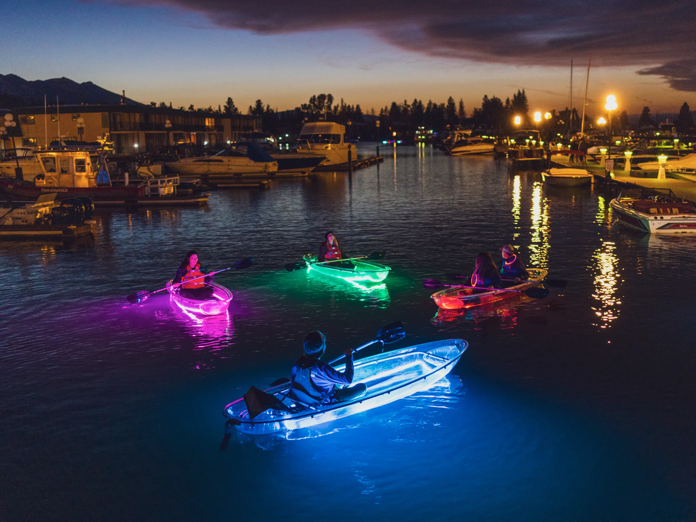 People in glowing kayaks on water at night near a marina.