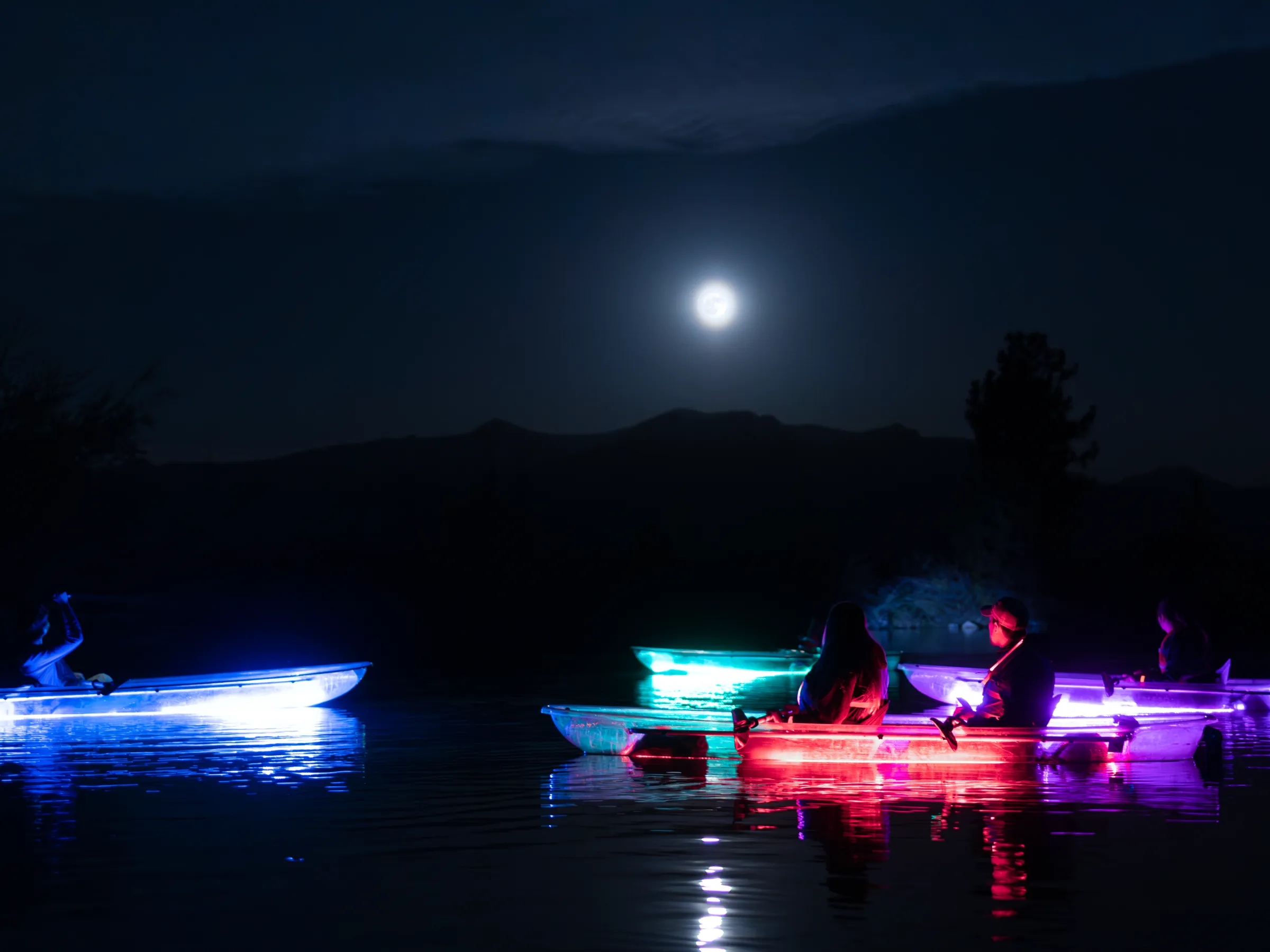 Boats with colorful lights on a lake at night under a full moon.