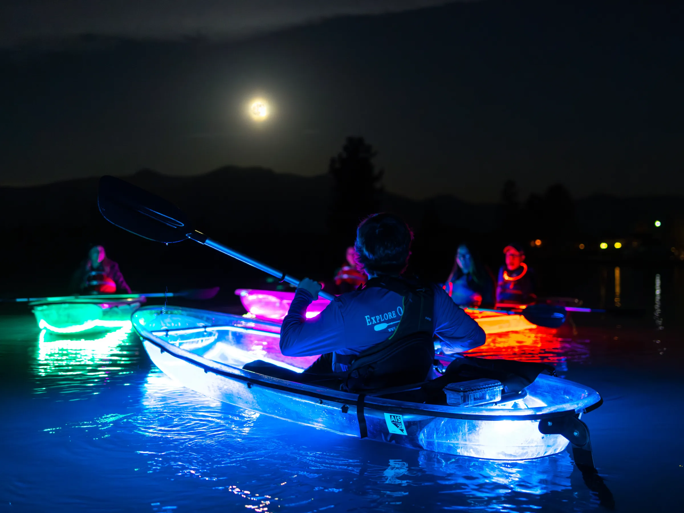 People in glowing kayaks paddle under a full moon at night.