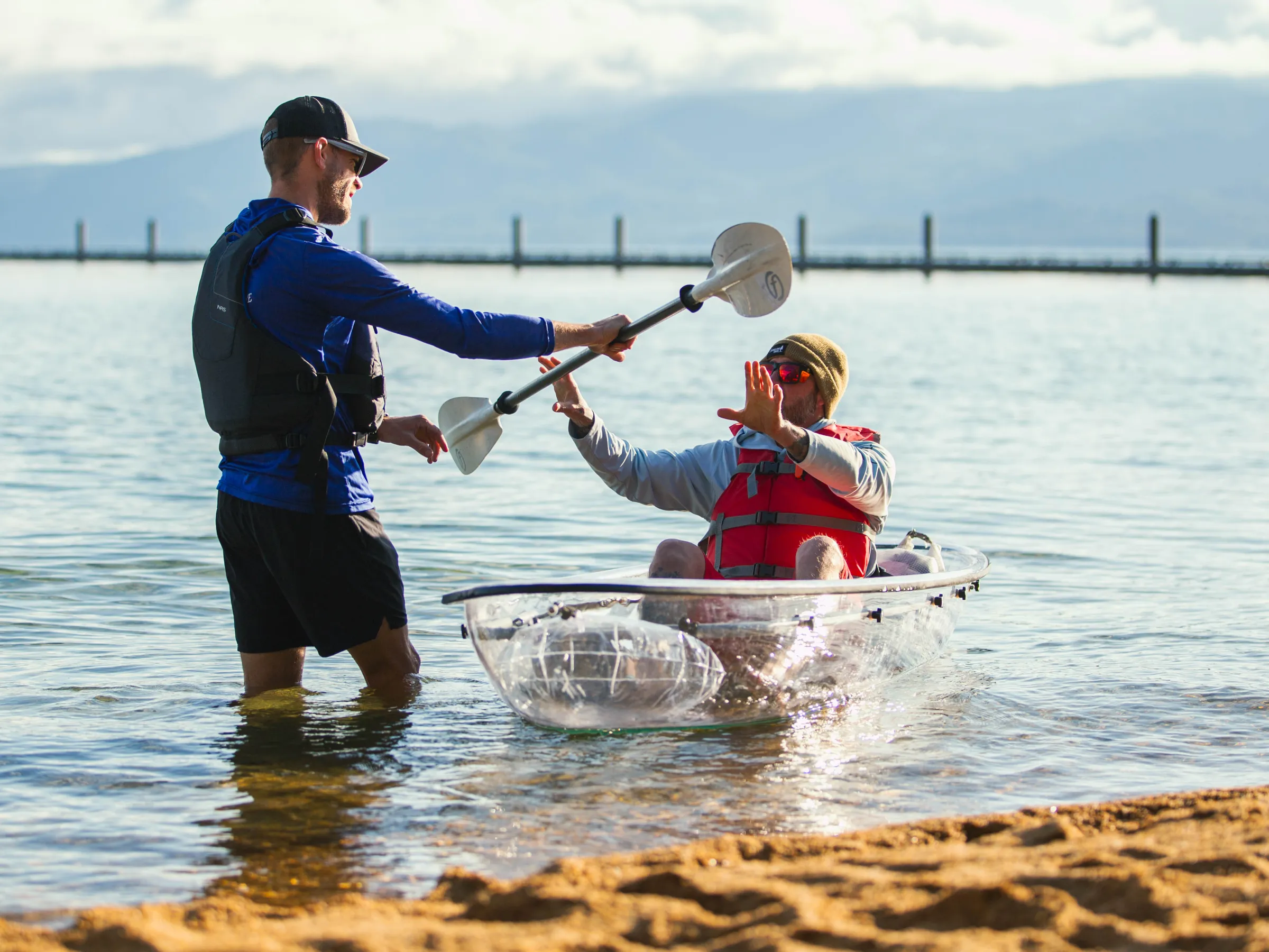 Two men by a clear kayak in shallow water; one standing, the other seated.