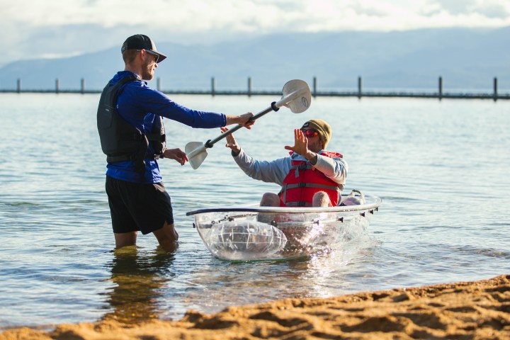 Two men by a clear kayak in shallow water; one standing, the other seated.