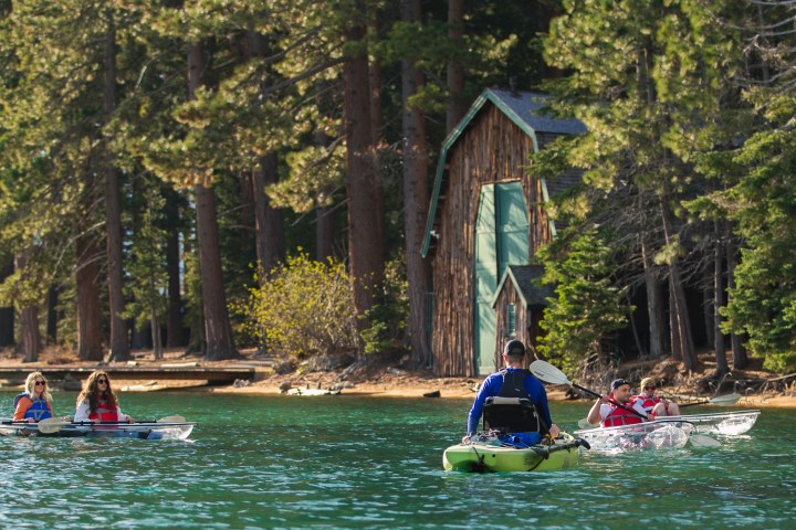 People kayaking on a lake with a wooden cabin and tall trees in the background.