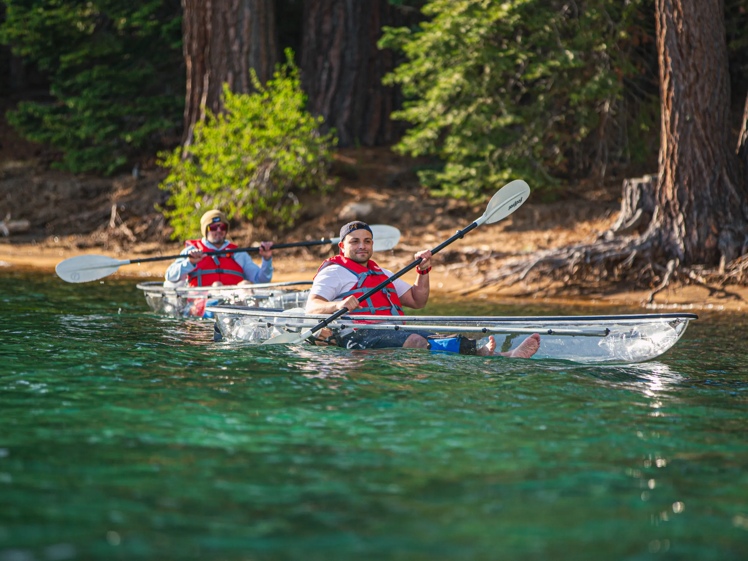 Two people kayaking in clear kayaks on a forested lake.