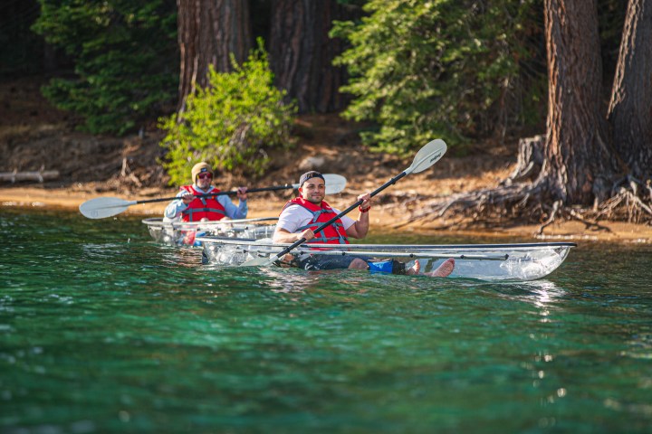 Two people kayaking in clear kayaks on a forested lake.