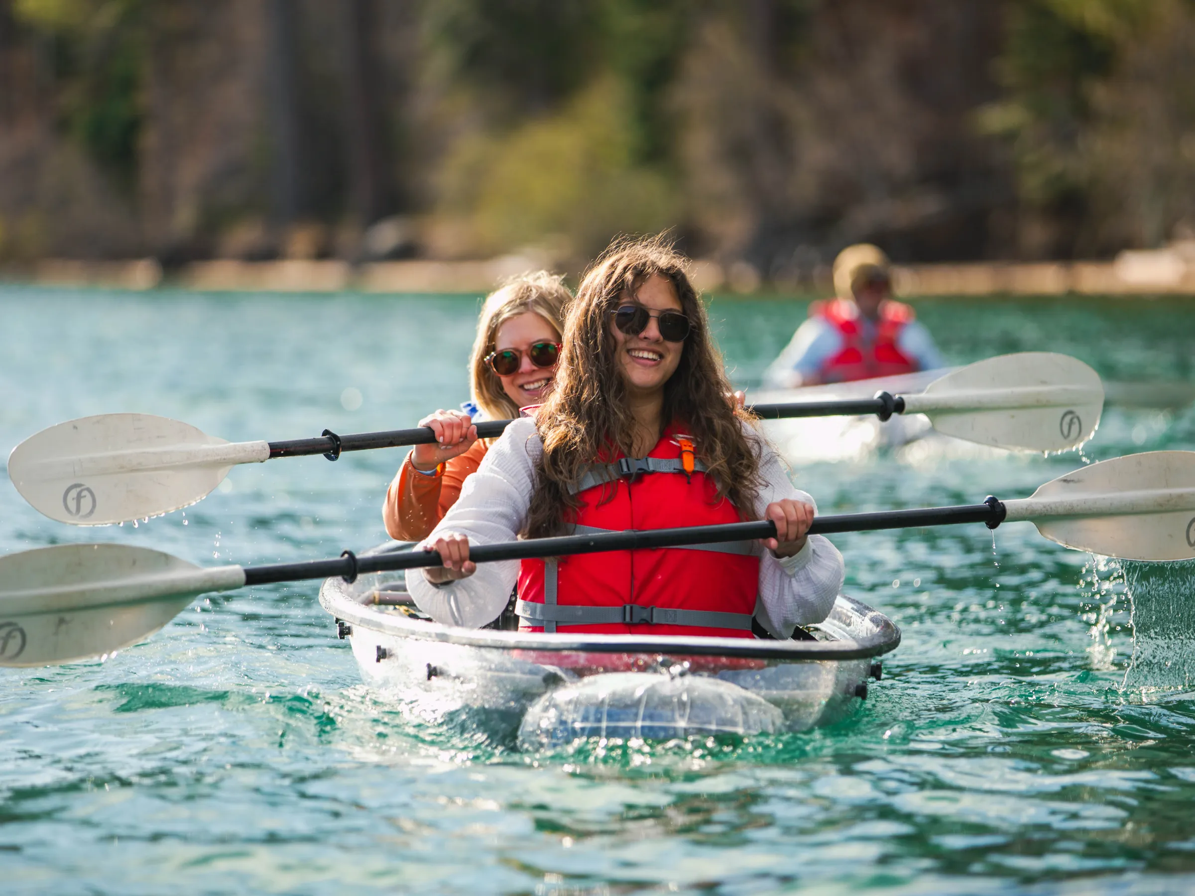 Two women kayaking on a lake with smiles, wearing red life vests.